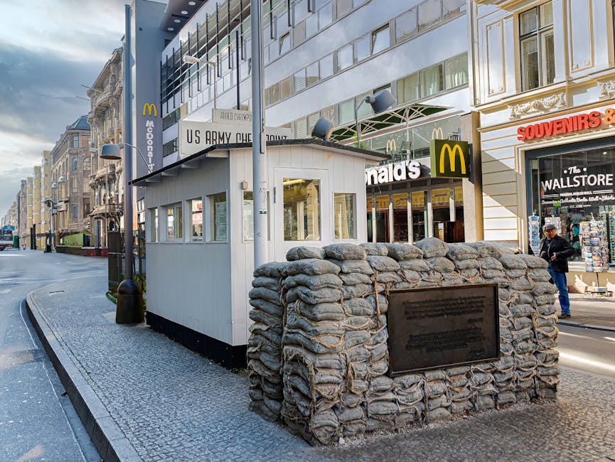 A small white guard booth with multiple windows is situated on a paved sidewalk in an urban environment, surrounded by a low, curved stone wall built from irregularly shaped stones. Attached to the st